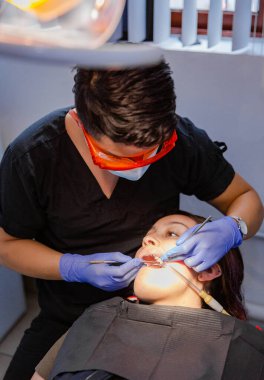 Diş hekimi muayenehanesindeki hasta diş beyazlatma ameliyatı oluyor. Diş hekiminde yüz maskesi, eldiven ve koruyucu gözlüklü PPE var..