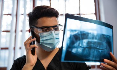 Dişçi kliniğinde bir radyografiyi inceliyor. Telefonda konuşuyor.