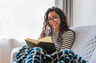 Mutlu genç kız oturma odasında bir kitap okuyor. Onun bacaklarında bir battaniye var.