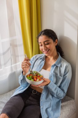 Genç esmer kadın gülümsüyor ve salata yiyor. O, odasında bir kanepede oturuyor.