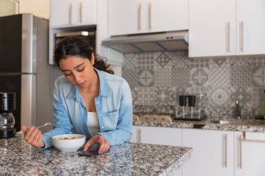 Genç esmer kadın mutfağında salata yiyor. Telefonunu kullanıyor.