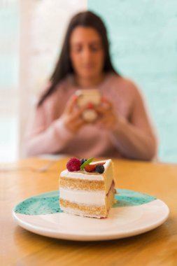 Masanın üzerindeki kırmızı çilekli pastayı kapat. Arka planda telefonunu kullanan bir kadın var.