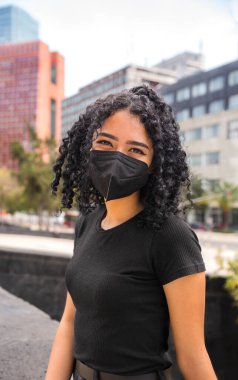 Kıvırcık saçlı genç kadın oturuyor. Siyah bir maske takıyor.