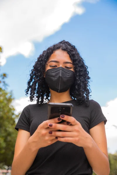 Kıvırcık saçlı genç kadın telefonunu kullanıyor, yüzünde siyah bir maske var.
