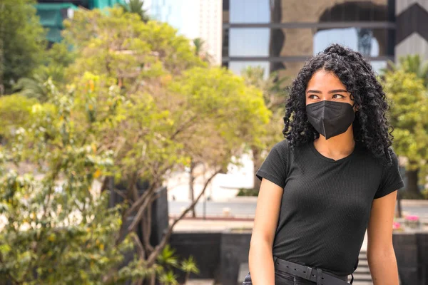 Kıvırcık saçlı genç kadın oturuyor. Siyah bir maske takıyor.
