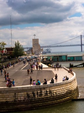 Lizbon 'daki Tagus Nehri kıyısındaki işlek rıhtım. Uzakta Keşifler Anıtı var. Turistler öğle yemeklerini yiyip geziyorlar. Vasco de Gama köprüsü arka planda görülebiliyor.