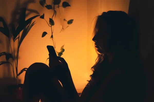 Gece boyunca evde uzun saçlı güzel bir genç kadın. Spot ışıkları altında akıllı telefon kullanan karanlık bir odada. Siyah arka plan ve sarı ışık. İçerideki portre.