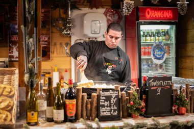 Perpignan, Fransa 'da aydınlatılmış bir pazar yerinde, tezgahın arkasında geleneksel yemek hazırlayan ya da pişiren bir satıcıyı özenle yakalayan samimi bir fotoğraf. Görüntü, sezonun odaklanmış çalışmalarını ve gastronomisini vurguluyor..