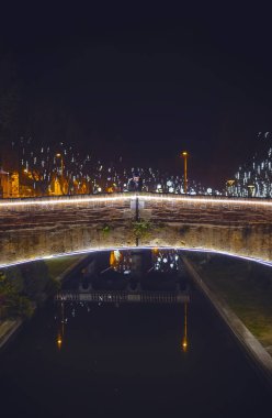 Fransa 'nın Perpignan şehrinde, bir kanalın üzerindeki tarihi bir köprüde, gece yarısı sicim ışıklarıyla aydınlanan bir kadının dikey çekimi. Romantik şehir manzarasını ve şenlikli atmosferi yakalar..