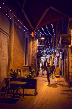 Fransa 'nın Perpignan kentindeki dar ve aydınlık bir caddede (sokak arasında) gece yürürken çekilen bir fotoğraf. Sahnede tuğla duvarlar, dükkan tabelaları ve tepende asılı duran şenlikli iplik ışıkları yer alıyor..
