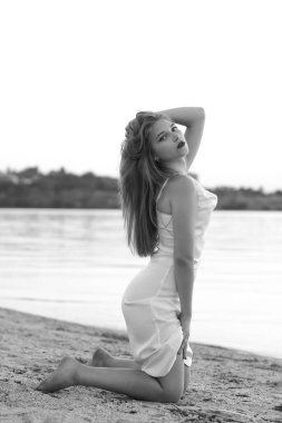 Güzel kız sahilde dizlerinin üzerinde oturur ve kameraya bakar. Nehre yakın, beyaz ipek elbise giymiş. Siyah beyaz resim