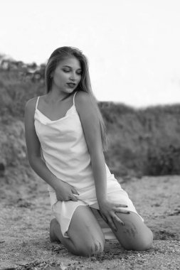 Güzel bir kız kumsalda dizlerinin üzerinde oturuyor ve elinde bir elbise tutuyor. Nehre yakın, beyaz ipek elbise giymiş. Siyah beyaz resim 