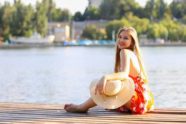 One beautiful girl sits on a wooden bridge and looks into camera smiling. She is near river side, dressed into orange flower pattern dress. It is summer sunset. She has got her hat  in one hand