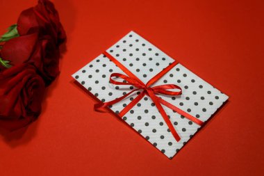 İçinde hediye zarfı, kırmızı kurdele, sertifika ve davetiye olan üç kırmızı gül. El işi konsepti. Kırmızı arka planda kırmızı. Metin için kopyalanma alanı. Düğün, tören konsepti, tatiller ve sürprizler.  