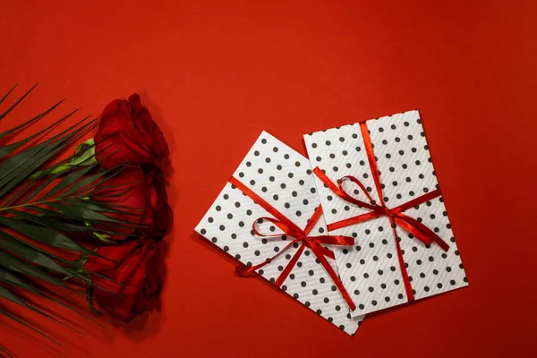 3 red roses with 2 gifts envelopes, red ribbons, certificates, invitation card inside. Craft concept. Red on the red background. Copyspace for text. Weddig, ceremony concept, holidays and surprises.