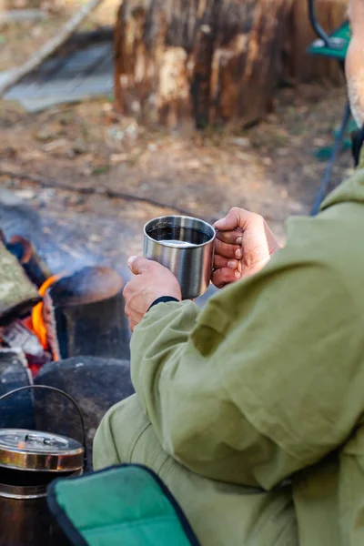 Young man travelling alone, drinking warm tea near the bonfire from metal mug. Concept of active tourism, camping in the forecast, trekking and hiking during vacations. Close up