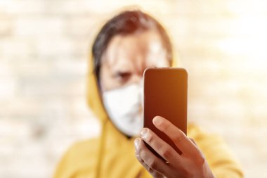 Maskeli genç adam sarı kapüşonlu akıllı telefonuna bakıyor. Parlak ışık.