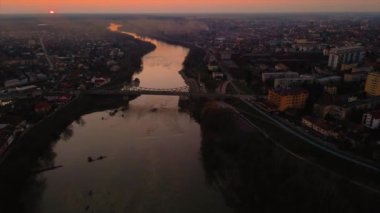 Gün batımında nehir ve köprü uzanan bir şehir manzarasının havadan videosu. Kamera yukarı doğru kayarken dron soldan sağa hareket ediyor ve nehri odaklıyor. Nehirli bir şehrin üzerinde gün batımının robot görüntüleri..