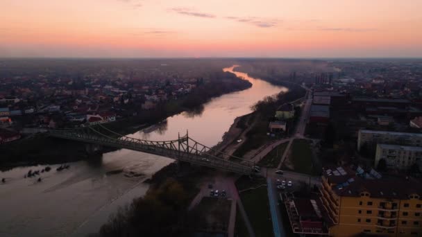 川のある街並みと夕日の広がる橋の空中ビデオ ドローン離れて橋から飛んでいると高度を拾うも 右に曲がっている 日没時に川と街のドローン映像 — ストック動画