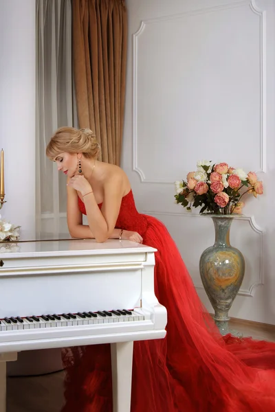 Donna in abito lungo rosso appoggiata al pianoforte bianco — Foto Stock