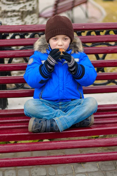 Ragazzo in giacca blu mangia torta con semi di papavero su una panchina — Foto Stock