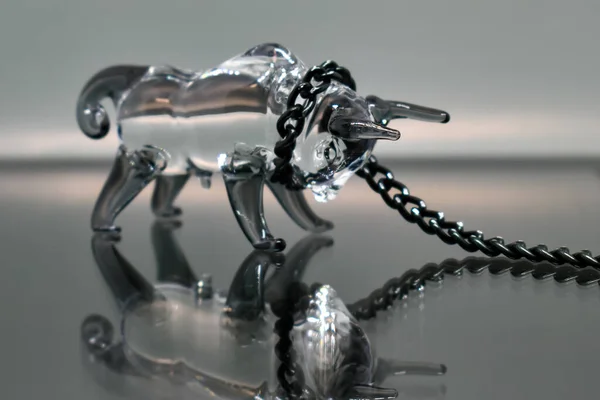 A glass figurine of a bull with a black metal chain around its neck stands against a mirrored background, looking at its reflection. Bull on a chain. Selective focus. Symbol of the year 2021