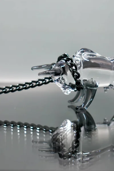 A glass figurine of a bull with a black metal chain around its neck stands against a mirrored background, looking at its reflection. Bull on a chain. Selective focus. Symbol of the year 2021