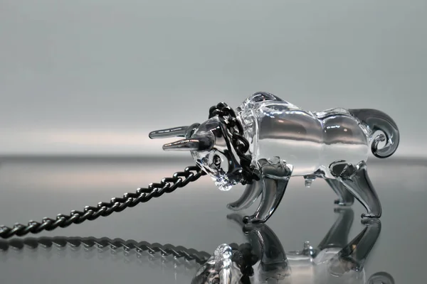 A glass figurine of a bull with a black metal chain around its neck stands against a mirrored background, looking at its reflection. Bull on a chain. Selective focus. Symbol of the year 2021