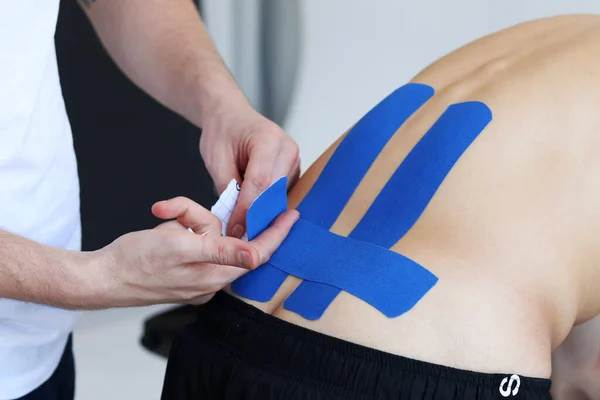 Fizyoterapist, erkek hastanın beline mavi kinesio bandı uyguluyor. Kineziyoloji, fizik tedavi, rehabilitasyon konsepti.