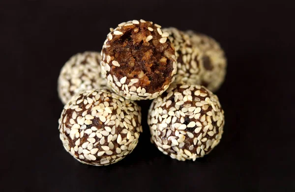 Vegan raw sweet balls on a black background. Natural sweets from nuts and dried fruits in coconut, cocoa and sesame seeds, close-up. Sugar-free and gluten-free sweets, raw food desserts