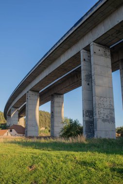 Büyük beton otoyol köprüsünün altında sıcak bir akşam güneşi altında büyük sütunlar fotoğraflandı. Manzarada modern inşaat mühendisliği ve ulaşım altyapısı.