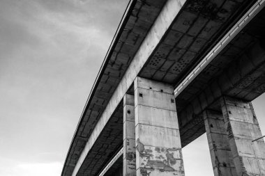 Gökyüzüne karşı kocaman sütunları olan bir otoyol köprüsünün alçak açılı monokrom fotoğrafı. Güç, geometri, yapı ve endüstriyel tasarımı vurgulayan soyut perspektif.