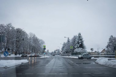 Soğuk bir kış gününde, yolun iki tarafında da karla kaplı ağaçlar olan geniş bir şehir caddesi. Bu görüntü, kış ulaşımı, şehir altyapısı, karayolu güvenliği, gidiş geliş ve kentsel alanlarda mevsimsel hava koşulları gibi konular için idealdir..
