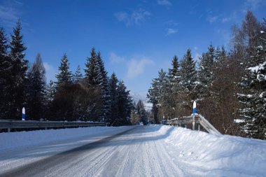Açık mavi gökyüzü altındaki yoğun bir ormanın içinden geçen taze karla kaplı sakin bir kış yolu. Görüntü, kış seyahatleri, soğuk hava koşulları, yol güvenliği, doğa ve kışın huzurlu kırsal manzaraları temsil ediyor..