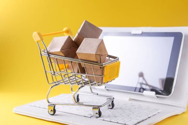 Kutuları olan alışveriş arabası, sarı arka planda beyaz dizüstü bilgisayar. İnternet Çevrimiçi alışveriş kavramı.