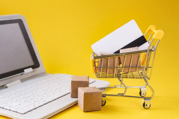 Shopping cart with boxes, credit card, white laptop on yellow background. Internet Online shopping concept.