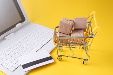 Kutulu alışveriş arabası, kredi kartı, sarı arka planda beyaz laptop. İnternet Çevrimiçi alışveriş kavramı.