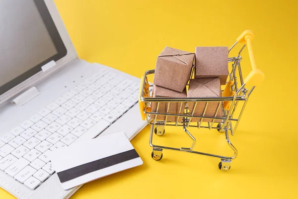 Winkelwagen met vakken, creditcard, witte laptop op gele achtergrond. Internet Online winkelen concept. — Stockfoto