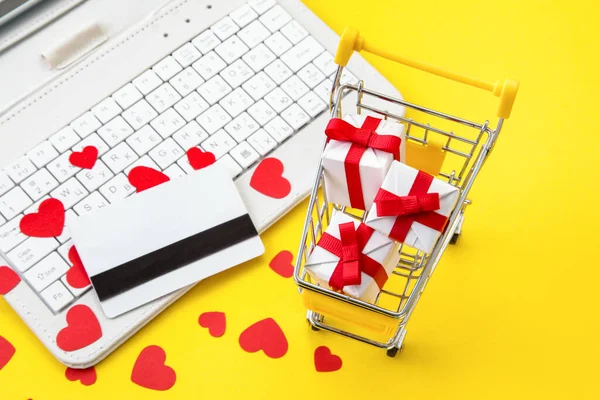 Online winkelen tijdens de vakantie. Valentijnsdag cadeaus bestellen. Winkelwagen met geschenkverpakking, creditcard, hartjes en witte laptop op gele achtergrond. — Stockfoto