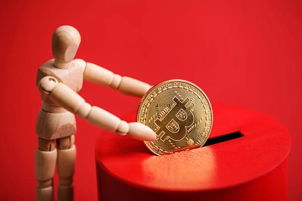 Wooden man puts gold coin with bitcoin symbol into slot of red donation box. Concept crypto currency. Selective focus — Stock Photo, Image