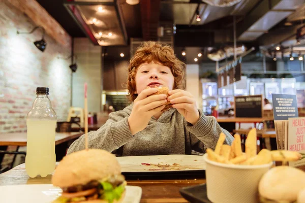 Hungrig pojke äter hamburgare i restaurang — Stockfoto