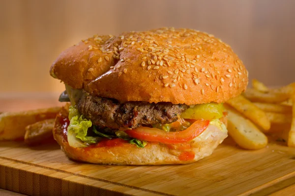 Burger lezat dengan keju yang meleleh dan patty daging sapi, selada, tomat, bawang, roti wijen tebal berdiri di atas meja kayu — Stok Foto
