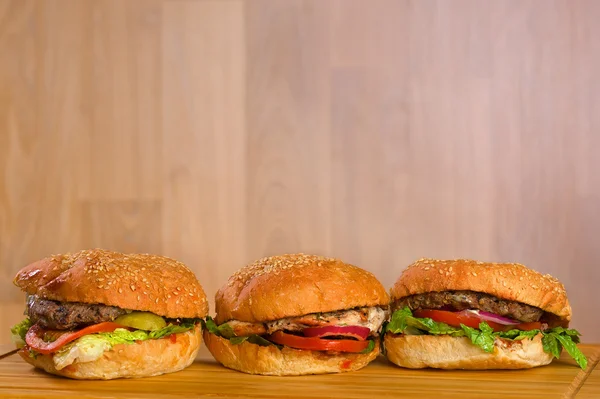 Tres sabrosas hamburguesas con queso derretido y carne molida suculenta gruesa empanada, pollo, lechuga, tomate, cebolla, pan de sésamo de pie en una fila sobre una mesa de madera —  Fotos de Stock