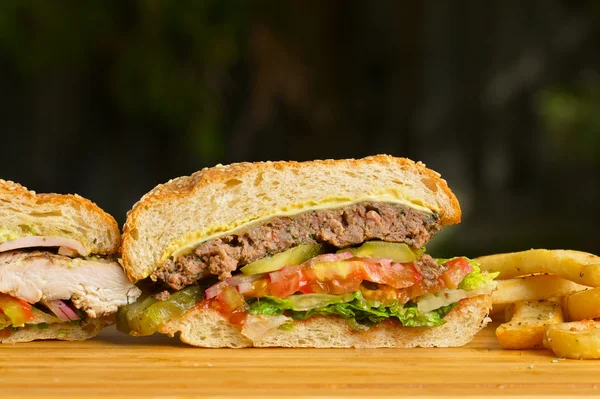 Doi burgeri tăiați cu brânză topită și carne de vită suculentă groasă măcinată, pui la grătar, salată verde, roșii, ceapă, chifle de susan în picioare pe masă de lemn — Fotografie, imagine de stoc