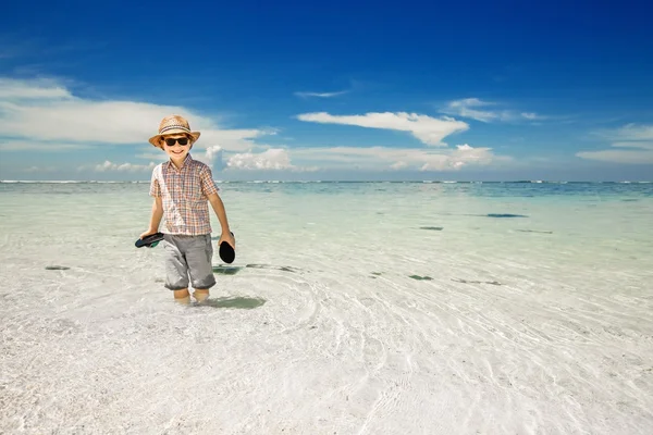 Szczęśliwy młody chłopiec przebywa w wody oceanu na pięknej plaży na sobie kapelusz i okulary. — Zdjęcie stockowe