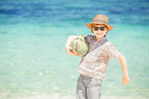 Szczęśliwy młody chłopak przebywający na piękny ocean plaża na sobie kapelusz i okulary gospodarstwa kokosowe w jego heands — Zdjęcie stockowe