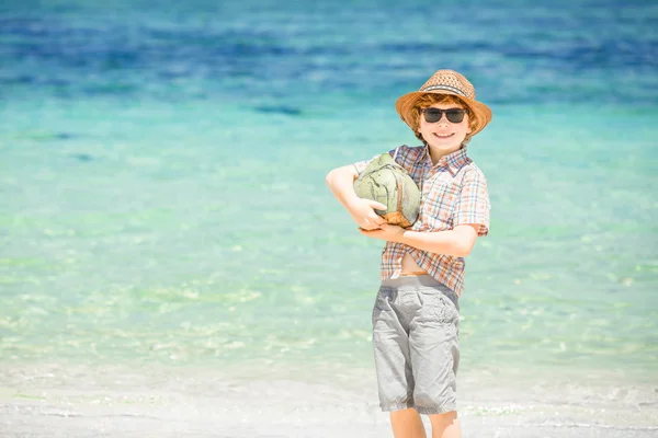 Щасливий молодий хлопець залишається на красивому океанському пляжі в капелюсі і сонцезахисних окулярах, тримаючи кокос в його плечах — стокове фото