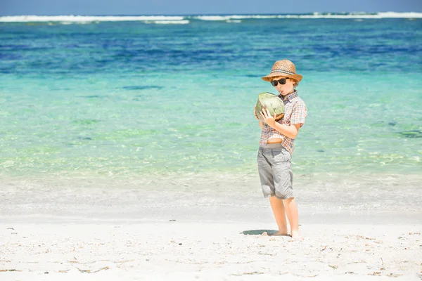 Szczęśliwy młody chłopak przebywający na piękny ocean plaża na sobie kapelusz i okulary gospodarstwa kokosowe w jego heands — Zdjęcie stockowe