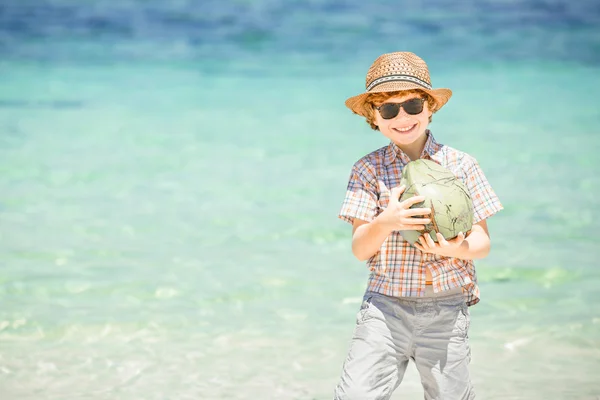 Un băiat fericit care stă pe plaja frumoasă a oceanului purtând pălărie și ochelari de soare care țin nucă de cocos în hățile sale — Fotografie, imagine de stoc