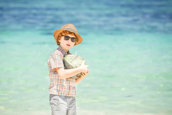 Щасливий молодий хлопець залишається на красивому океанському пляжі в капелюсі і сонцезахисних окулярах, тримаючи кокос в його плечах — стокове фото